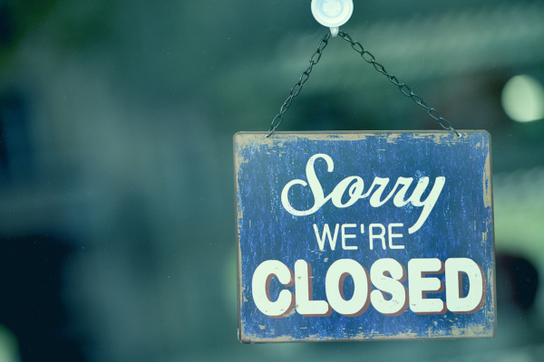 A blue and white sign hanging on a glass door reads Sorry We’re Closed.