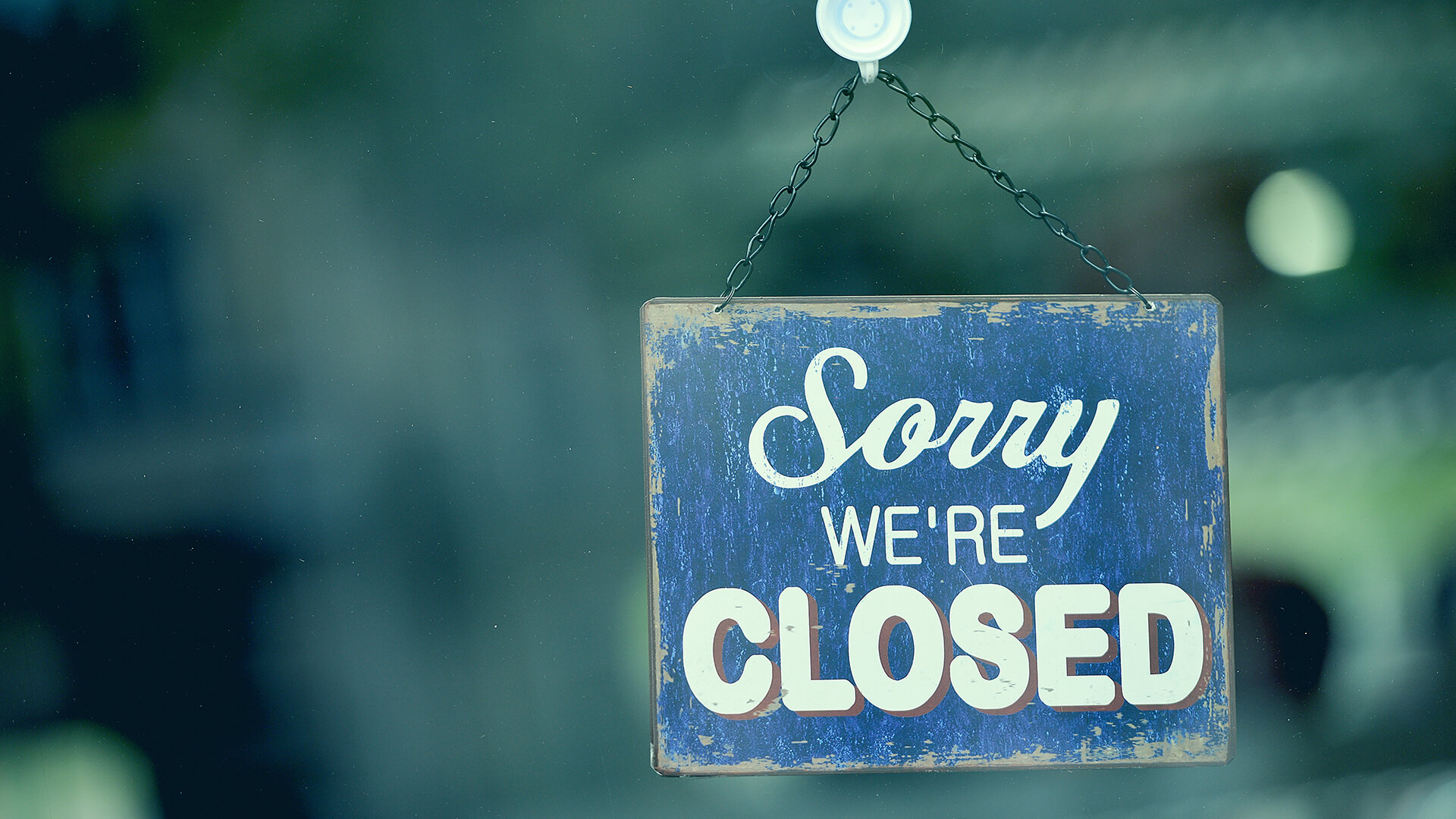 A blue and white sign hanging on a glass door reads Sorry We’re Closed.