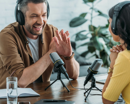 Two people wearing headphones sit at a table with microphones and recording equipment, chatting. A notebook, pen, and glass of water are on the table, with a plant visible in the background.