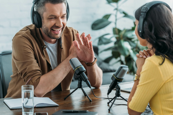 Two people wearing headphones sit at a table with microphones and recording equipment, chatting. A notebook, pen, and glass of water are on the table, with a plant visible in the background.