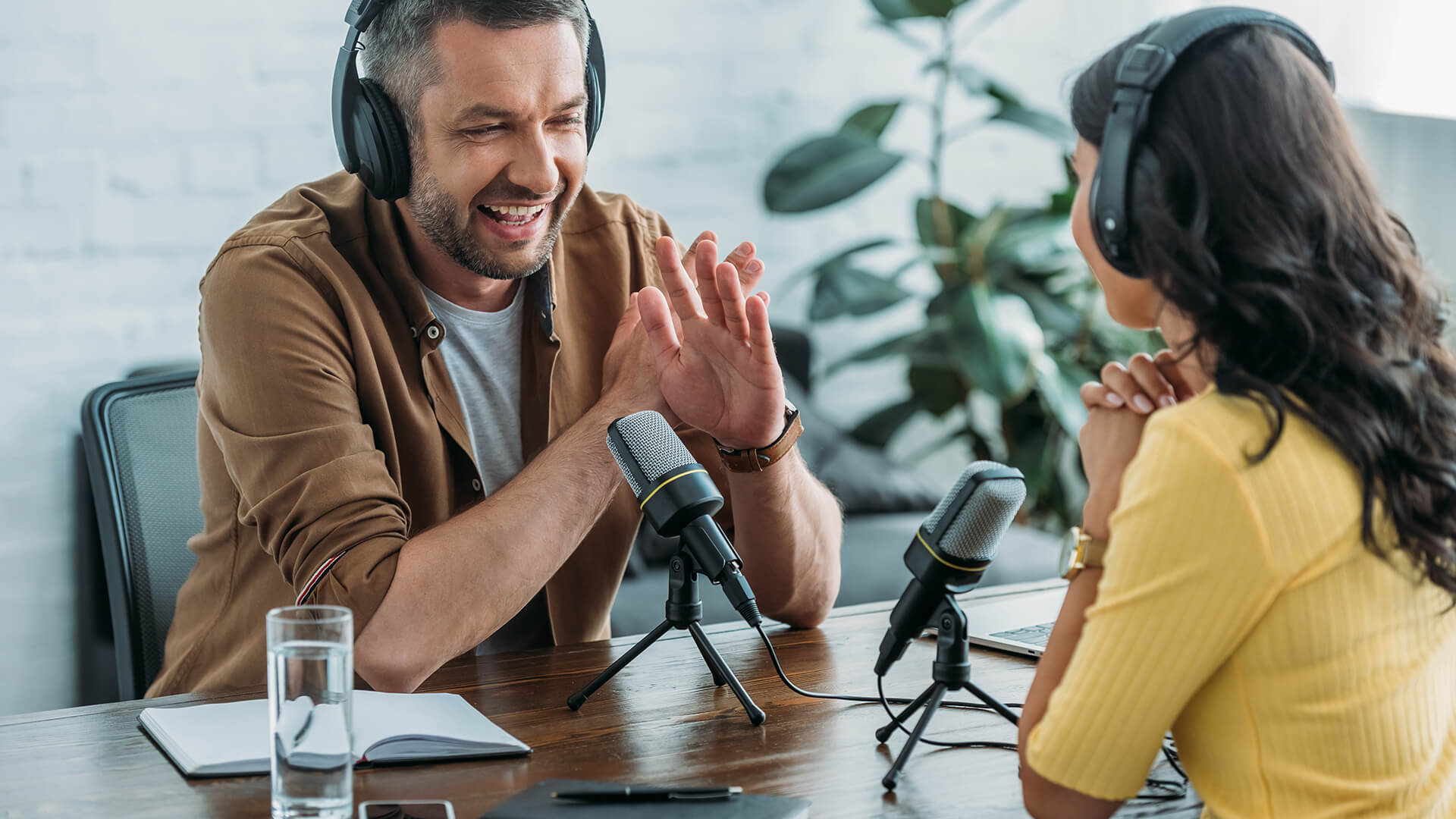 blog-podcast-day Two people wearing headphones sit at a table with microphones and recording equipment, chatting. A notebook, pen, and glass of water are on the table, with a plant visible in the background.