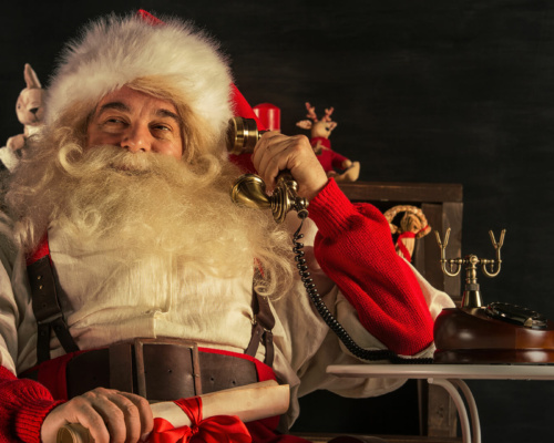 Father Christmas sits in a chair, holding a scroll tied with a red ribbon whilst talking on an old-fashioned telephone for Christmas on hold messages.