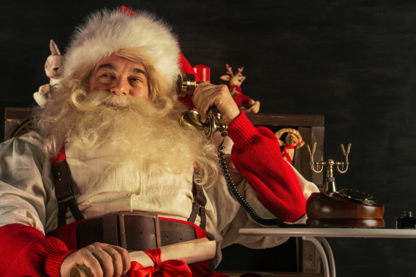Father Christmas sits in a chair, holding a scroll tied with a red ribbon whilst talking on an old-fashioned telephone for Christmas on hold messages.