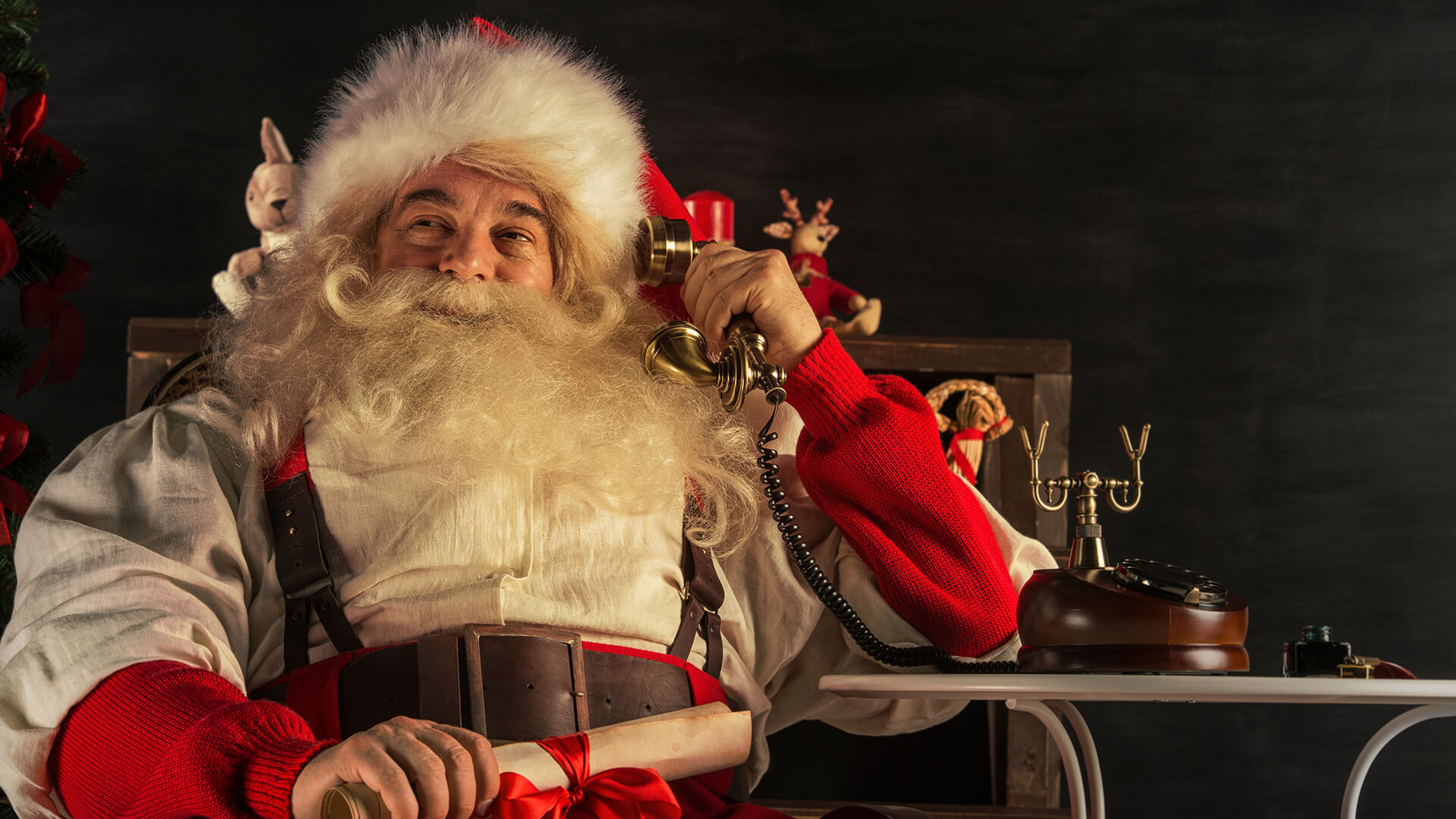 Father Christmas sits in a chair, holding a scroll tied with a red ribbon whilst talking on an old-fashioned telephone for Christmas on hold messages.