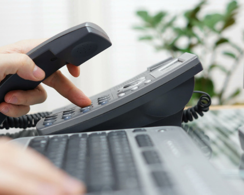 A person dials a number on an office landline phone, possibly using multi-site messaging, whilst holding the receiver; a computer keyboard and monitor are visible in the foreground.