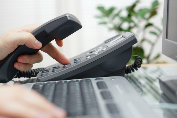 A person dials a number on an office landline phone, possibly using multi-site messaging, whilst holding the receiver; a computer keyboard and monitor are visible in the foreground.