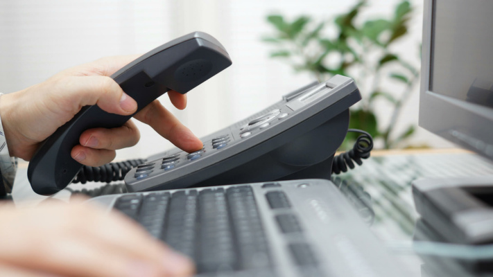 blog-scaling-phone A person dials a number on an office landline phone, possibly using multi-site messaging, whilst holding the receiver; a computer keyboard and monitor are visible in the foreground.
