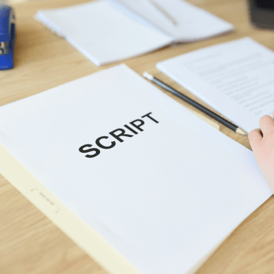 A hand rests on a table beside a stack of papers marked SCRIPT in bold letters, with additional documents and a pen seen in the background.
