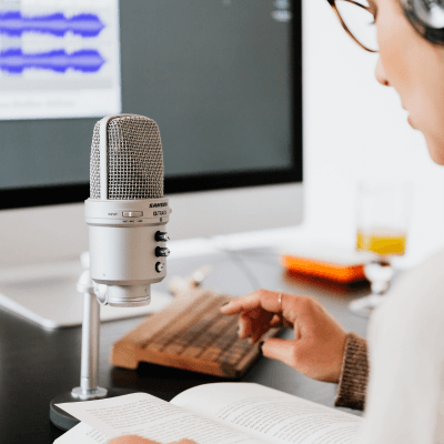 A person sits at a desk with an open book, typing on a keyboard beside a microphone, while a computer monitor in the background displays audio waveforms.