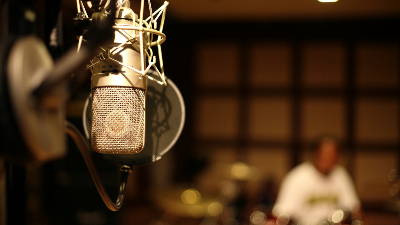 Close-up of a professional microphone and pop filter in a recording studio, with a person blurred in the background.