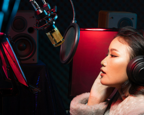 A person in headphones and a fur coat is recording or speaking into a microphone in a studio, with audio equipment and loudspeakers visible in the background.