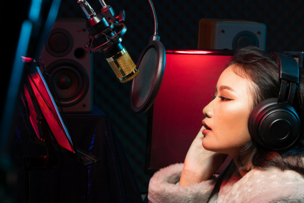 A person in headphones and a fur coat is recording or speaking into a microphone in a studio, with audio equipment and loudspeakers visible in the background.