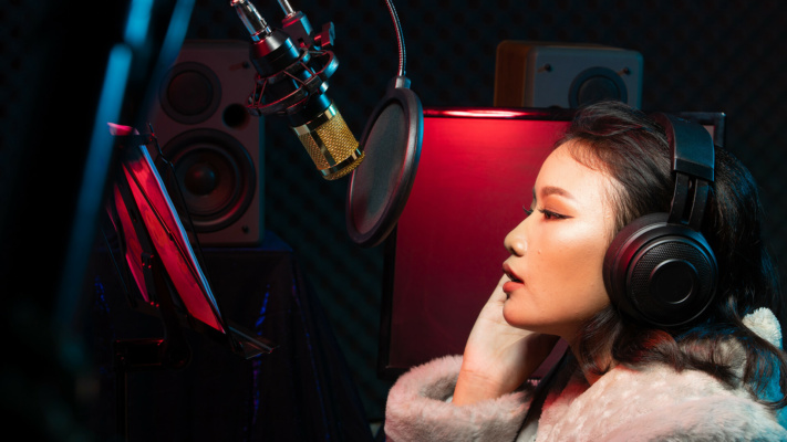A person in headphones and a fur coat is recording or speaking into a microphone in a studio, with audio equipment and loudspeakers visible in the background.