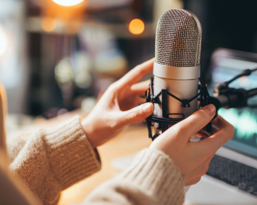 Individual adjusting a large condenser microphone on a desk, with a laptop showing audio waveforms in the background, indicating a professional voiceover or podcasting set-up.