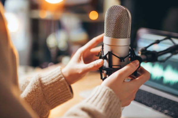 Individual adjusting a large condenser microphone on a desk, with a laptop showing audio waveforms in the background, indicating a professional voiceover or podcasting set-up.