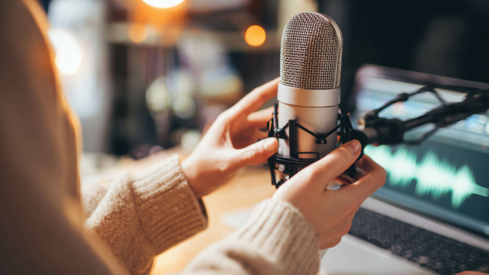 blog-voice-over Individual adjusting a large condenser microphone on a desk, with a laptop showing audio waveforms in the background, indicating a professional voiceover or podcasting set-up.