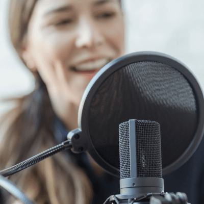 A woman speaks into a microphone with a pop filter in front of her, suggesting she is recording audio or taking part in a podcast.