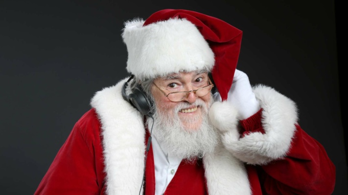 A person dressed as Santa, smiling with spectacles and wearing headphones, adjusts the hat with one hand against a dark background.