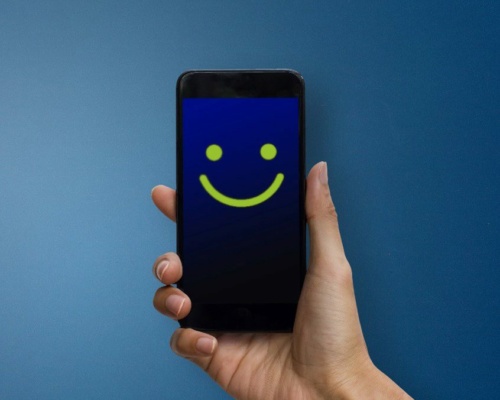 customer-experience-thumb A hand holds a mobile phone displaying a yellow smiley face on a blue screen against a blue background.