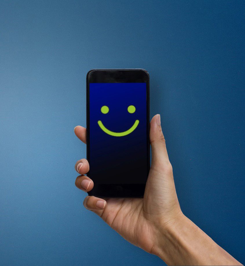 A hand holds a mobile phone displaying a yellow smiley face on a blue screen against a blue background.