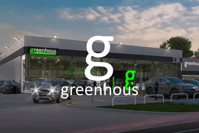 A modern car dealership building labelled Greenhous sits next to a Hobbycraft shop, with several parked cars in front beneath a partly cloudy sky.