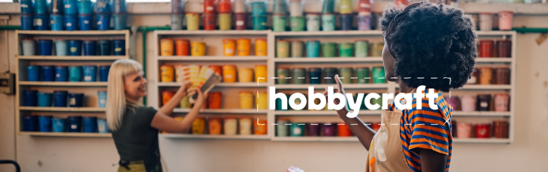 Two individuals in a craft shop surrounded by shelves of colourful paint tins. One person is reaching for a tin while the other observes. The word Hobbycraft is overlaid on the image.