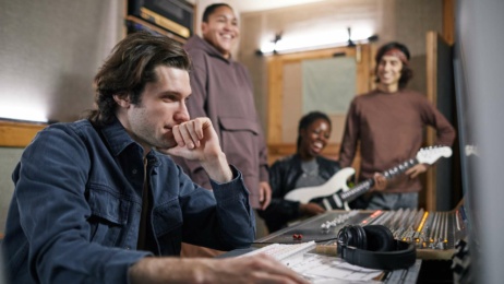 A man sits at an audio mixing desk in a recording studio while three people, one holding an electric guitar, stand and smile in the background.