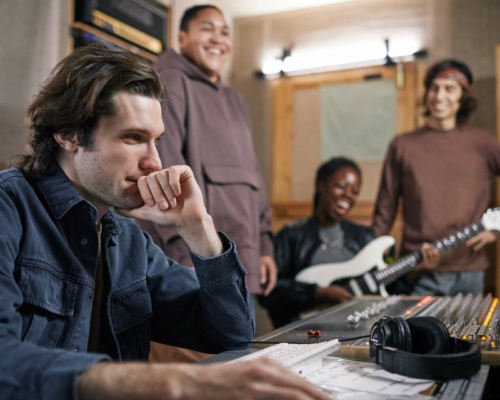 A man sits at an audio mixing desk in a recording studio while three people, one holding an electric guitar, stand and smile in the background.