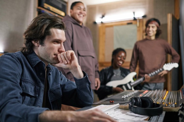 A man sits at an audio mixing desk in a recording studio while three people, one holding an electric guitar, stand and smile in the background.