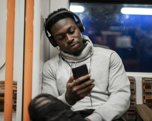 A person sits on a train, wearing headphones and looking at a smartphone, likely attuned to some on-hold marketing trend. Dressed in a grey hoodie and ripped jeans, they seem lost in their own digital world amidst the bustling commute.