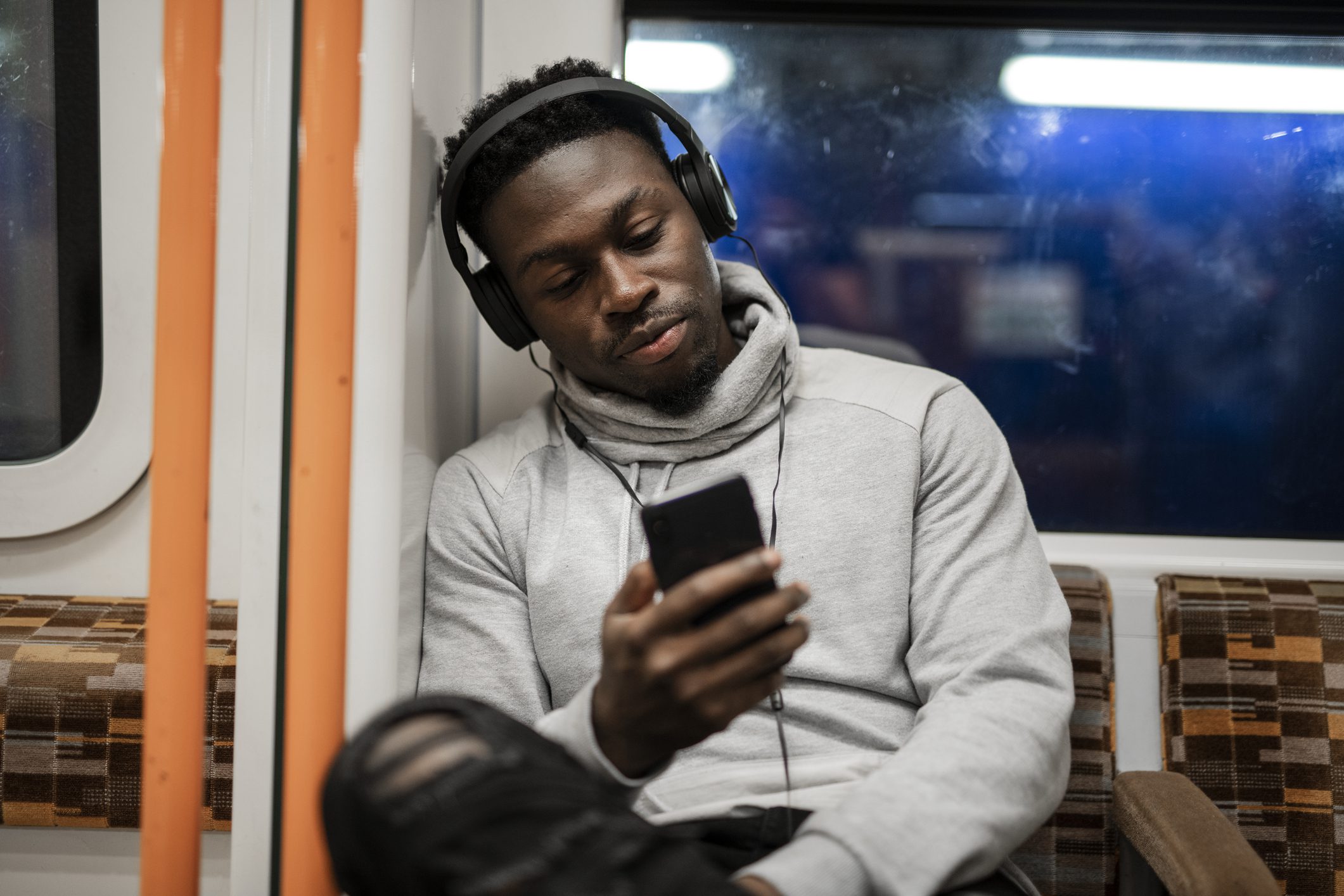 A person sits on a train, wearing headphones and looking at a smartphone, likely attuned to some on-hold marketing trend. Dressed in a grey hoodie and ripped jeans, they seem lost in their own digital world amidst the bustling commute.