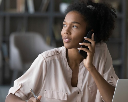Dreamy young african american woman holding telephone call. A woman is at her desk in an office, talking on the phone and taking notes while her laptop hums quietly beside her. As she navigates through the conversation, you can almost hear the soothing hold messages that punctuate this busy work environment.