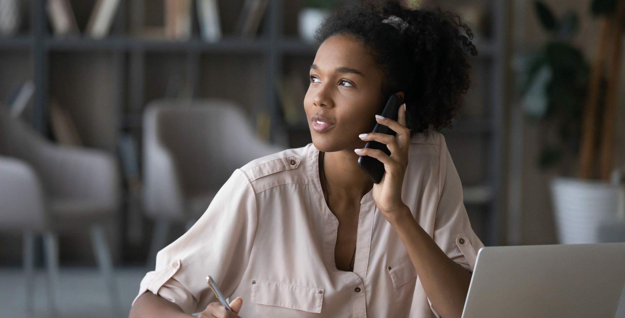 A woman is at her desk in an office, talking on the phone and taking notes while her laptop hums quietly beside her. As she navigates through the conversation, you can almost hear the soothing hold messages that punctuate this busy work environment.