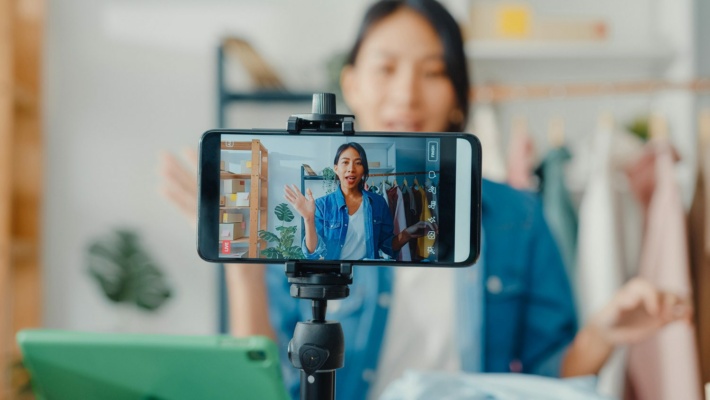 A person records a video with a smartphone on a tripod, embracing the art of video marketing. A clothes rail and green tablet enhance the scene in the background.