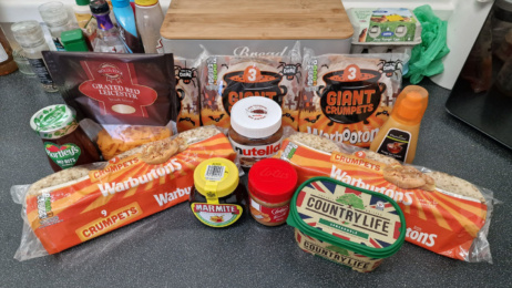 An assortment from Onhold Studio featuring a variety of crumpets, spreads, cheese, butter, and sauces graces the kitchen counter. The selection includes Warburtons crumpets, Nutella, Marmite, Lotus Biscoff, and Country Life butter.