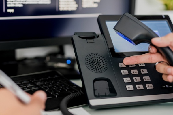 A person holds the handset of a desk phone, with a hand on the keypad, as gentle hold music plays in the background. Nearby, someone writes on paper, and a computer screen is partially visible.