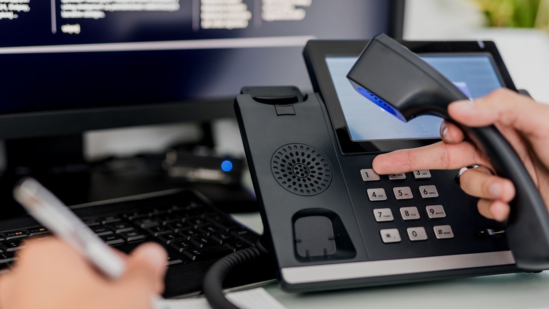 A person holds the handset of a desk phone, with a hand on the keypad, as gentle hold music plays in the background. Nearby, someone writes on paper, and a computer screen is partially visible.