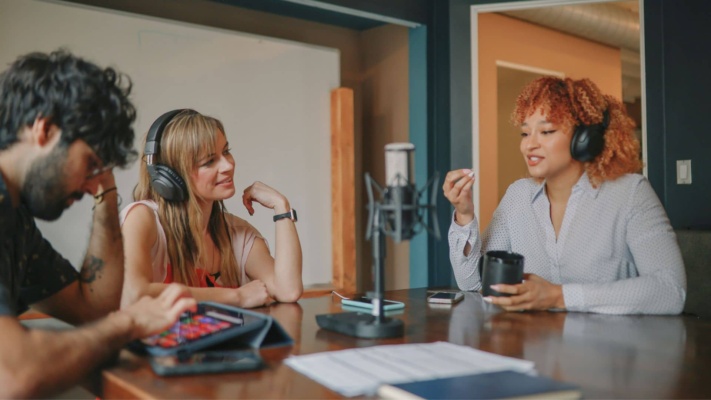 Three people wearing headphones are sitting around a table with a microphone, engaged in conversation. One person is holding a mug. A tablet and notepad are on the table.