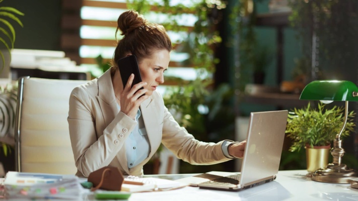 A woman in an office is on the phone while using a laptop. She is surrounded by plants and office stationery.