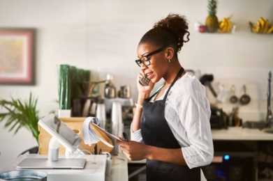 A woman wearing an apron discusses booking opportunities on the telephone while holding papers at a kitchen counter.