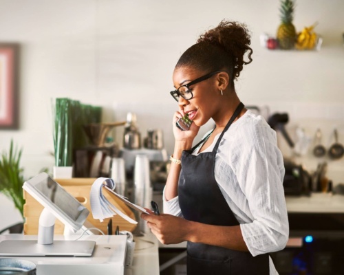 restaurant A woman wearing an apron discusses booking opportunities on the telephone while holding papers at a kitchen counter.