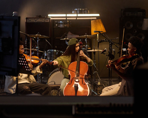 Three musicians play string instruments in a dimly lit studio, with drums, speakers, and music stands in the background.