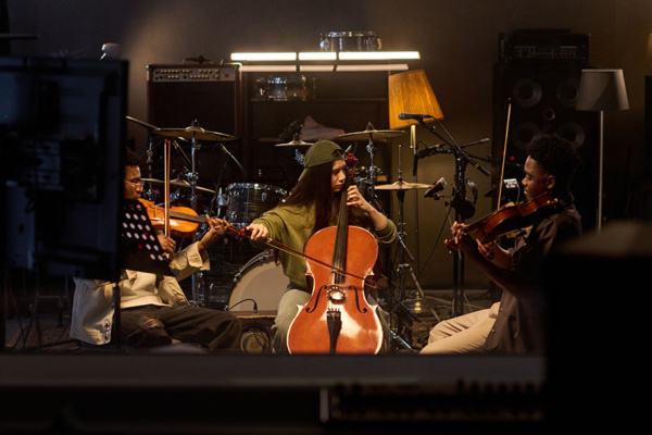 Three musicians play string instruments in a dimly lit studio, with drums, speakers, and music stands in the background.
