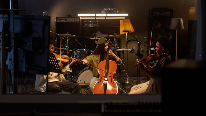 Three musicians play string instruments in a dimly lit studio, with drums, speakers, and music stands in the background.