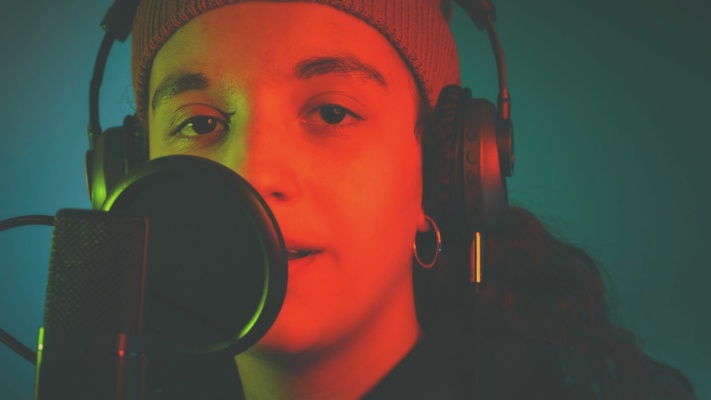 A person wearing headphones and a knitted cap speaks into a microphone, the atmosphere enhanced by red and green lighting, embodying the art of professional voice recordings.