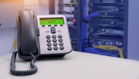 A cloud-based VoIP telephone sits on the desk in a server room, with blurred server racks and tangled cables forming the backdrop.