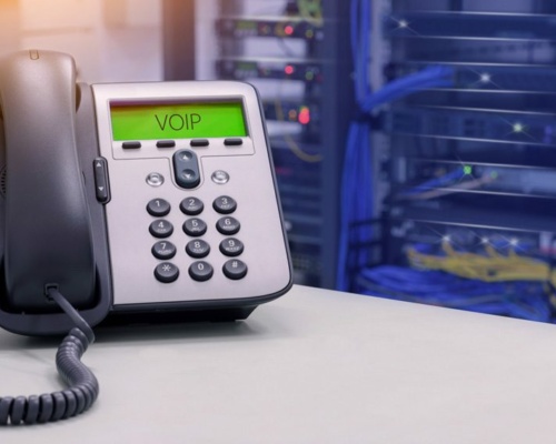 A cloud-based VoIP telephone sits on the desk in a server room, with blurred server racks and tangled cables forming the backdrop.
