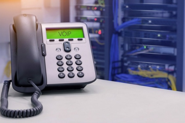 A cloud-based VoIP telephone sits on the desk in a server room, with blurred server racks and tangled cables forming the backdrop.