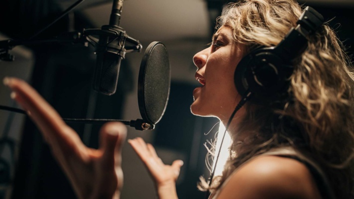 Person with curly hair wearing headphones, singing into a microphone in a recording studio.